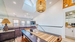 A spacious living room with a dark wood dining table, a woven rug, and a large window offering a view of the city. - OCTIPI Your SenSEAS - Kitty Hawk, Sea Dunes, Open All Year, Flex Stays, Pool, Clubhouse (Kitty Hawk)
