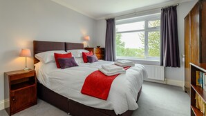 Una cama bien tendida con sábanas blancas y detalles rojos, una mesita de noche con una lámpara y vistas al exterior a través de una ventana con cortinas.