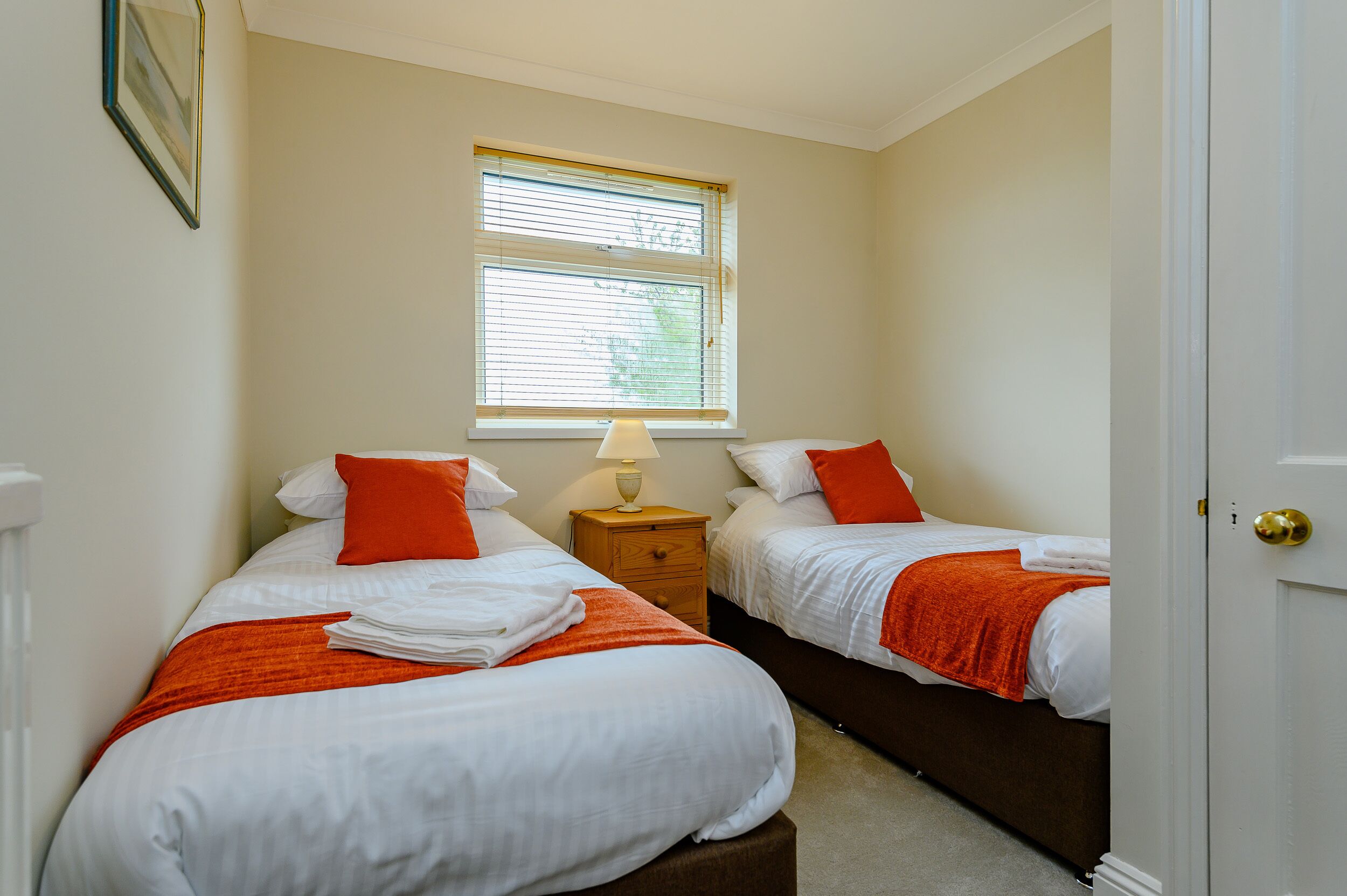 A bedroom with two beds, each with white bedding and orange accents, a nightstand with a lamp, and a window with blinds.