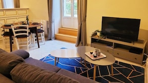 Een woonkamer met een bank, een eettafel met stoelen, een tv op een dressoir en een trap naar een buitenruimte.