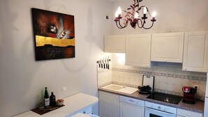 Een moderne keuken met witte kastjes, een aanrecht, een spoelbak, een fornuis en een kroonluchter. Er hangt een schilderij aan de muur en op het aanrecht staat een fles wijn.