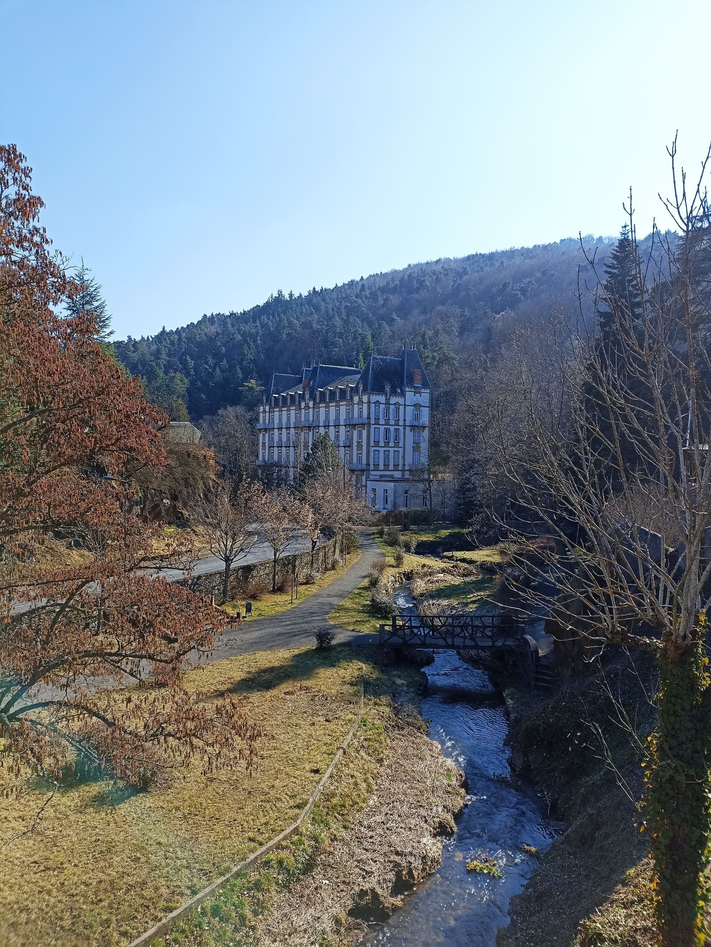 A large, historic building with multiple stories and a traditional architectural style, surrounded by a natural landscape with a river and trees.