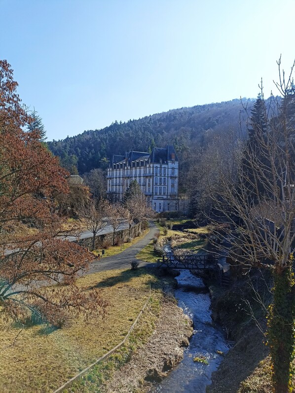 A large, historic building with multiple stories and a traditional architectural style, surrounded by a natural landscape with a river and trees.