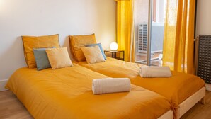 A double room with two beds, each with a yellow bedspread and multiple pillows. There is a small table with a lamp and a window with yellow curtains. - Champollion Appartment : A/C + Netflix + Garage (Albi)