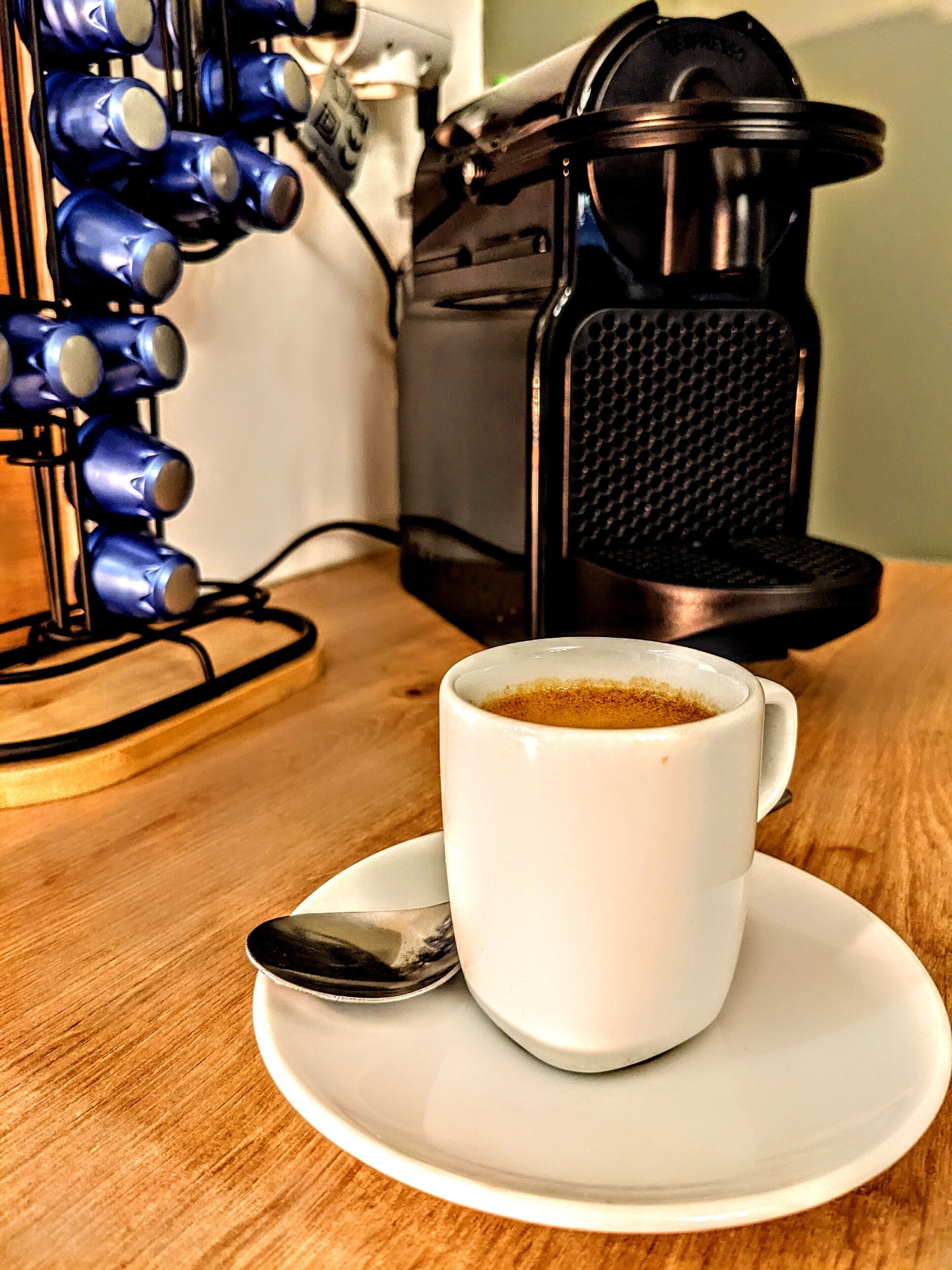 A coffee maker, a cup of coffee, and a spoon on a wooden table.