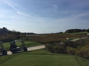 A golf course with a path leading to a clubhouse, a water body in the background, and a distant view of a house.