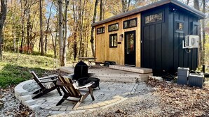 A wooden cabin with a porch, a picnic table, and a fire pit. - The Hemlock Tiny House (Logan)