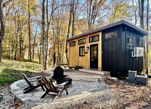 A cabin with a wooden exterior, a black door, and a small porch area with a bench and a fire pit. - The Hemlock Tiny House (Logan)