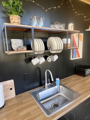 A modern kitchen with a stainless steel sink, wooden countertops, and a black backsplash. There are shelves above the sink holding dishes and mugs, and a plant on the left. - The Hemlock Tiny House (Logan)