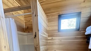 A wooden cabin bathroom with a shower, toilet, and a small window. - The Hemlock Tiny House (Logan)