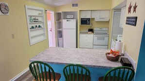 A compact kitchen with a central island, white cabinets, a microwave, a toaster, and a refrigerator. There are three green chairs around the island.