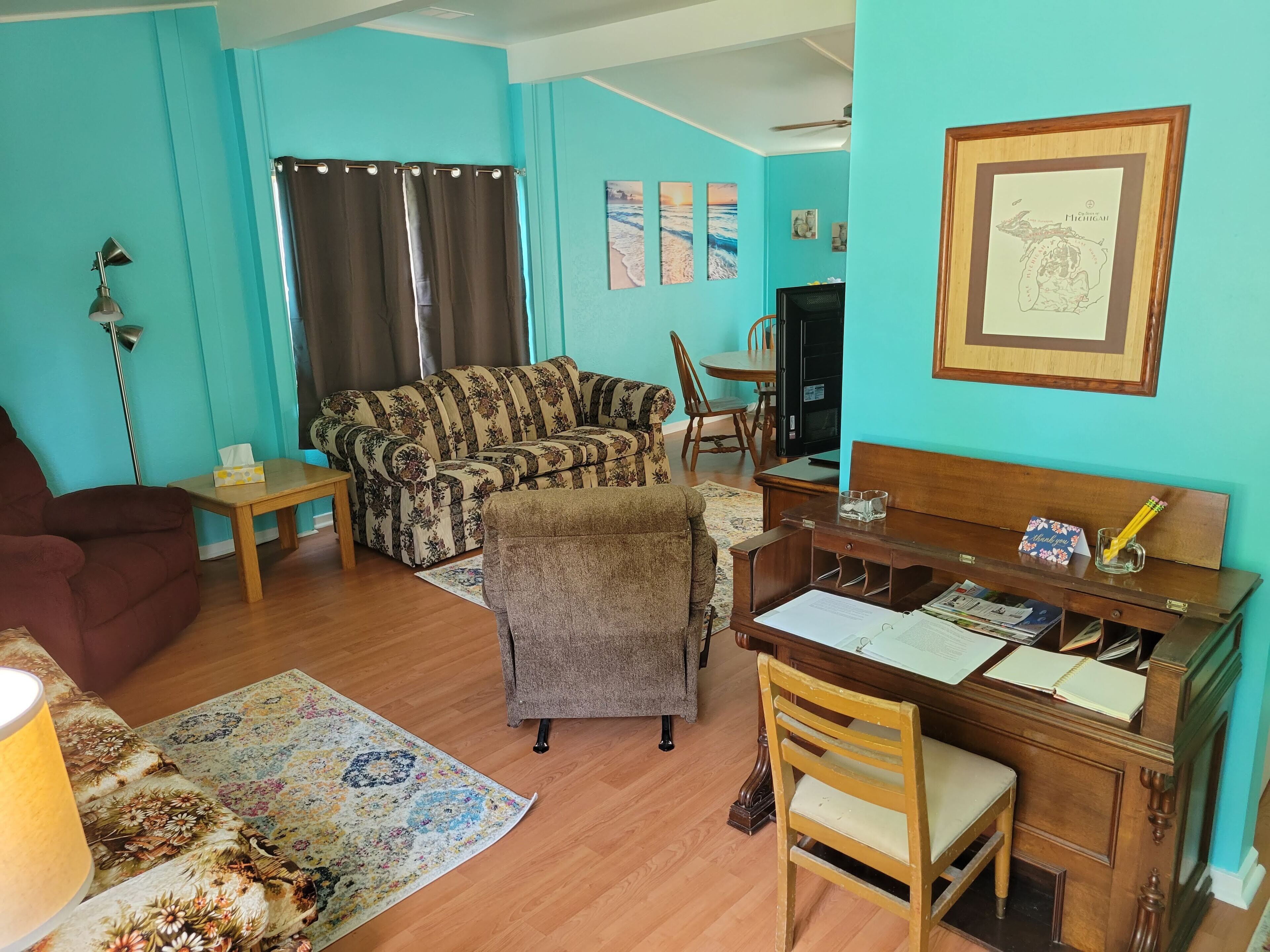 A living room with a sofa, armchair, coffee table, and a desk with a chair. There is a television, a lamp, and a framed picture on the wall.