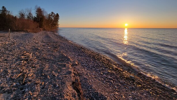 A pebbled beach with a sunset over the water, trees on the left, and a clear sky.
