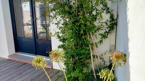 Ett trädäck med ett litet bord, en krukväxt och en blommande växt mot en vit vägg.