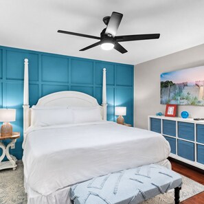 A bedroom with a bed, a ceiling fan, a nightstand, a lamp, a bedside table, a picture frame, a blue wall paneling, a window, and a painting on the wall.