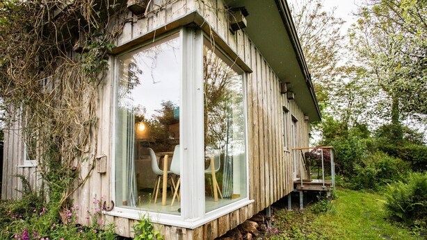 A wooden cabin with a glass front, a dining area with chairs, and a small outdoor deck.