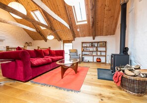 Un salon chaleureux avec un canapé rouge, une table basse en verre et une bibliothèque en bois. On aperçoit un poêle en bois noir à l’arrière-plan.
