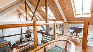 Una habitación estilo loft con techo alto, vigas de madera, barandilla de vidrio, chimenea, un sofá y una silla.