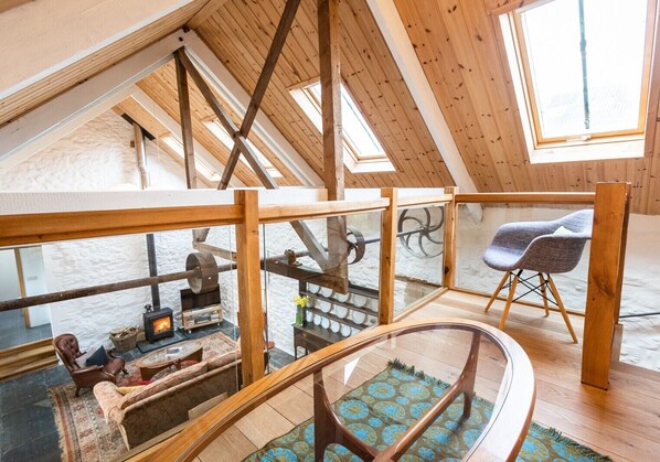 Une pièce de style « loft », avec un plafond haut, des poutres en bois, une rambarde en verre, une cheminée, un canapé et un fauteuil.