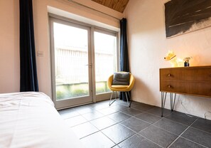 Une chambre avec un lit, une porte-fenêtre coulissante donnant sur l’extérieur, une chaise jaune, une commode en bois et une petite figurine de chien posée sur la commode.