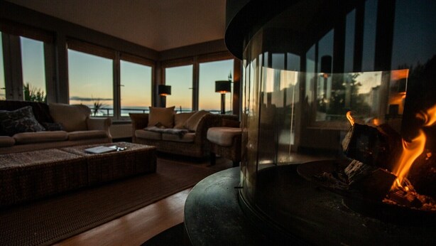 A modern living room with a large fireplace, comfortable seating, and large windows offering a view of the outdoors. - An exceptional place to gather on the sea shore (Newport)