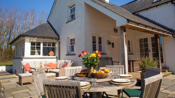 A patio area with a round wooden table set for dining, surrounded by wicker chairs. - A beautiful property for 6 with expansive lawns, minutes from the beach (Newport)