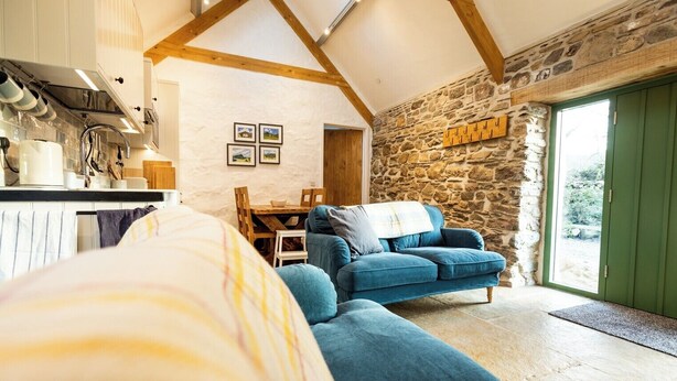 A cozy room with a stone wall, a blue sofa, a wooden dining table, and a kitchen area with white cabinets and a hanging pot rack.