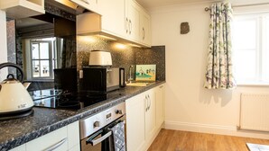 Une cuisine compacte équipée d’une cuisinière, d’une bouilloire, d’un four à micro-ondes et d’un petit four. Il y a une fenêtre avec des rideaux et un radiateur au mur.