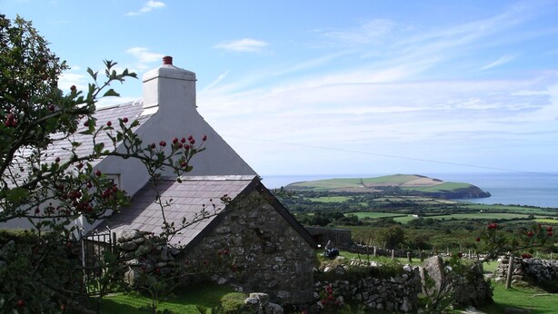 Une maison en pierre avec un toit blanc, entourée d’une zone herbeuse parsemée de baies rouges, et offrant une vue sur la mer et les collines en arrière-plan.