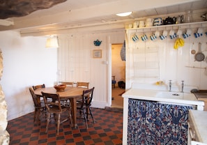 Un coin repas avec une table et des chaises en bois, un sol à carreaux, une porte blanche et une cuisine où les ustensiles sont suspendus.