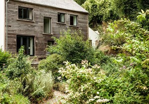 A modern two-story house with a sloped roof, surrounded by dense greenery and shrubs. - An incredible retreat on the coast for 10, with a beautiful garden (Cardigan)