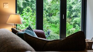 Ein gemütliches Zimmer mit einer Couch, einer Lampe und Blick auf einen üppigen Garten durch große Fenster.