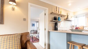 Un salon chaleureux avec un canapé en cuir marron et un coussin décoratif à motifs, qui donne sur une cuisine dotée d’un plan de travail bleu et de tabourets en bois.