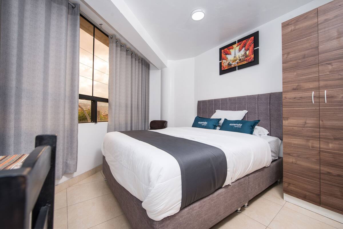 A bedroom with a large bed, a desk, a chair, a wardrobe, and a window with curtains.