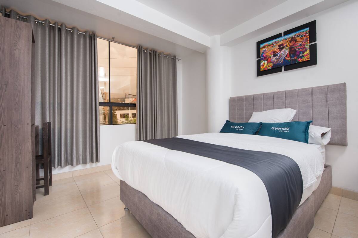 A modern hotel room with a large bed, grey curtains, a view of the city, and a colorful artwork on the wall.
