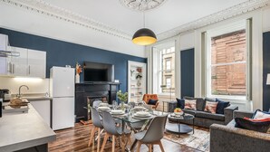 A modern kitchen with a dining area, a living room with a sofa and armchairs, and a large window with a view of a building.