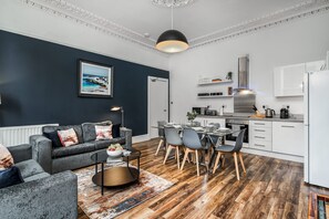 Uma sala de estar moderna com um sofá cinzento, uma mesa de jantar com cadeiras e uma área de cozinha com armários brancos e um exaustor de aço inoxidável.