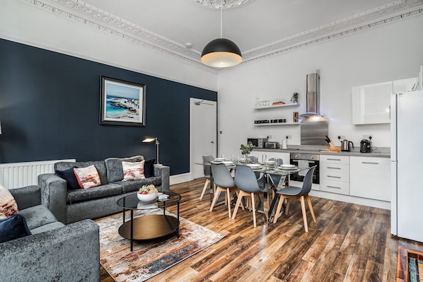 Un salon moderne avec un canapé gris, une table à manger avec des chaises et un coin cuisine doté de meubles blancs et d’une hotte en acier inoxydable.