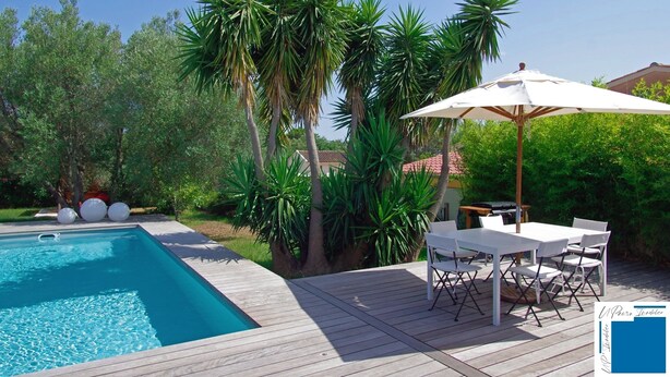 A swimming pool with a wooden deck, surrounded by lush greenery and a white umbrella.