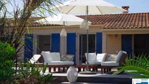 A patio with two white cushioned chairs, a round table, and a white umbrella providing shade.