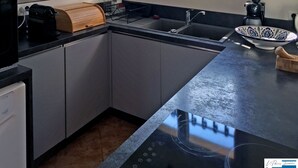 A modern kitchen with a black countertop, stainless steel sink, and a built-in oven. There are two wall-mounted coffee makers and a decorative bowl on the counter.