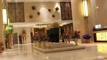 A hotel lobby with a reception desk, decorative plants, and a modern chandelier.