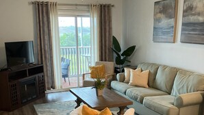 Ein Wohnzimmer mit einer Couch, einem Couchtisch, einem Fernseher und einem Balkon mit Vorhängen.