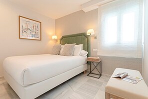 Habitación de hotel con una cama grande, una mesita de noche, otra mesita más pequeña, una lámpara de pared, un cuadro enmarco y una ventana con cortinas de encaje.