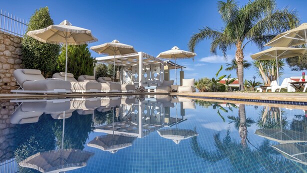 A poolside area with lounge chairs and umbrellas, surrounded by palm trees and a stone wall. - Azzuro Villa by Estia (Hersonissos)