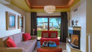 A living room with a beige sofa, red cushions, a red couch, and a fireplace. There are framed artworks on the walls and a view of the outdoors through large windows. - Azzuro Villa by Estia (Hersonissos)