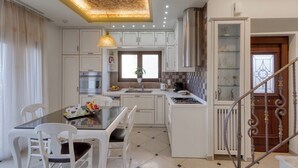 A modern kitchen with white cabinets, a dining table with chairs, a built-in oven, and a decorative ceiling light. - Sunshine VIlla by Estia (Hersonissos)