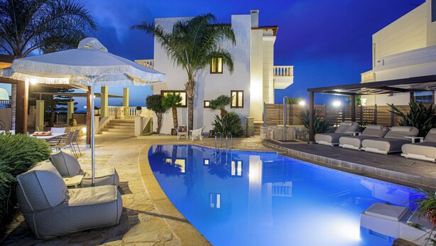 A swimming pool with lounge chairs and umbrellas, surrounded by a building with a white facade and a palm tree. - Sunshine VIlla by Estia (Hersonissos)
