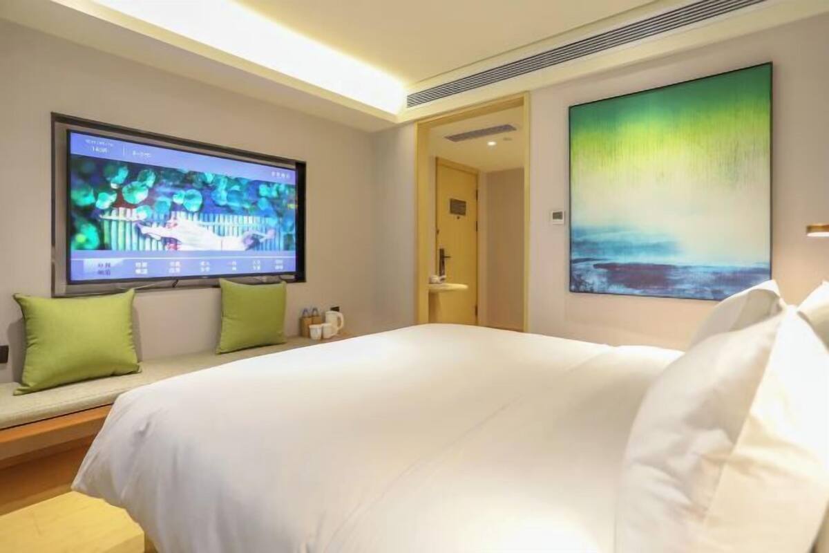 A hotel room with a large bed, a flat-screen TV mounted on the wall, a painting, and a bench with green cushions.