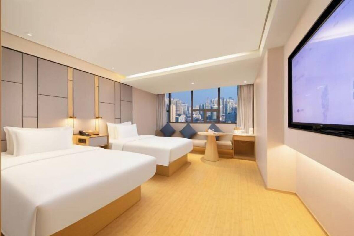 A modern hotel room with two beds, a sofa, a TV, and a city view.